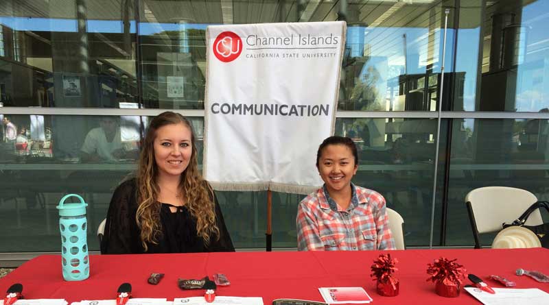 Two students sitting on a red table with a communication sign in the back